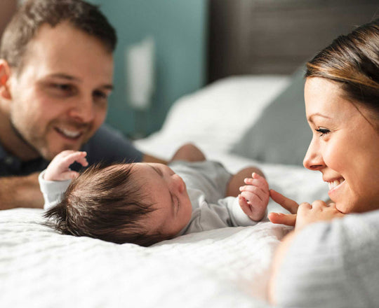 Ein Baby liegt auf einem Bett, während Mama und Papa daneben sitzen und es liebevoll ansehen