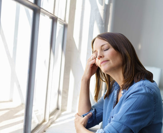 Frau leidet unter einer Frühjahrsmüdigkeit und steht vor dem Fenster, durch welches Sonne strahlt
