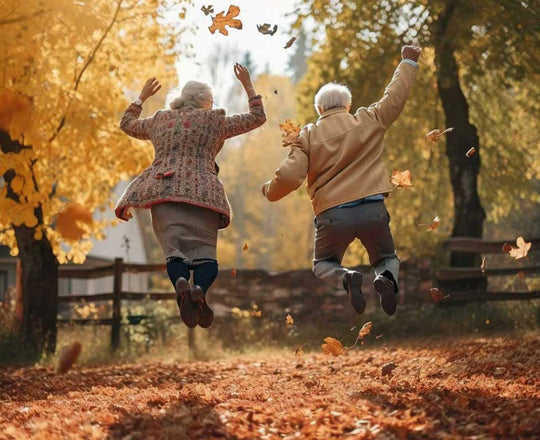 Ältere Frau und älterer Mann springen gemeinsam fröhlich in die Luft, umgeben von buntem Herbstlaub