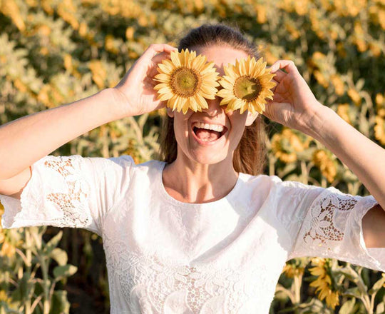 Eine Frau steht vor einem Sonnenblumenfeld und hält zwei Sonnenblumen vor ihre Augen