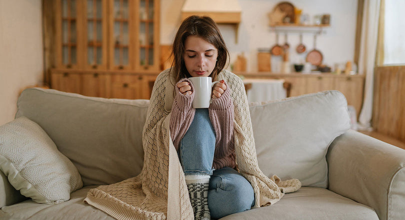 Eine Frau sitzt auf dem Sofa, eingewickelt in eine Decke, und hält eine Tasse Tee