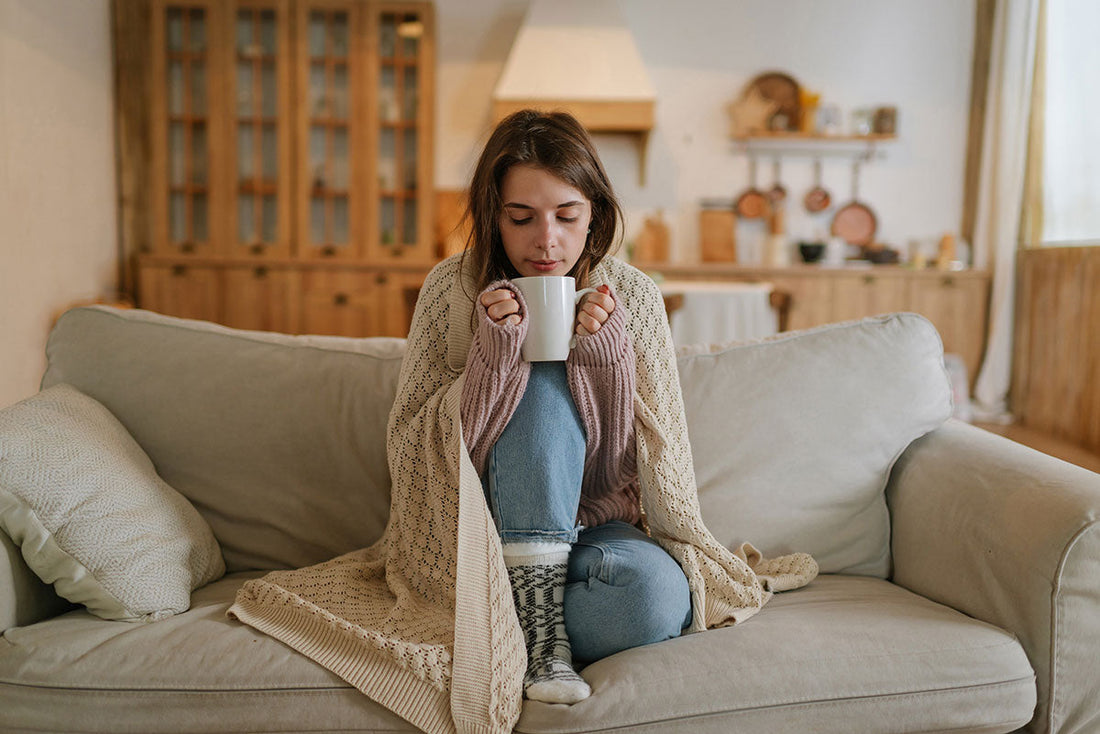 Eine Frau sitzt auf dem Sofa, eingewickelt in eine Decke, und hält eine Tasse Tee