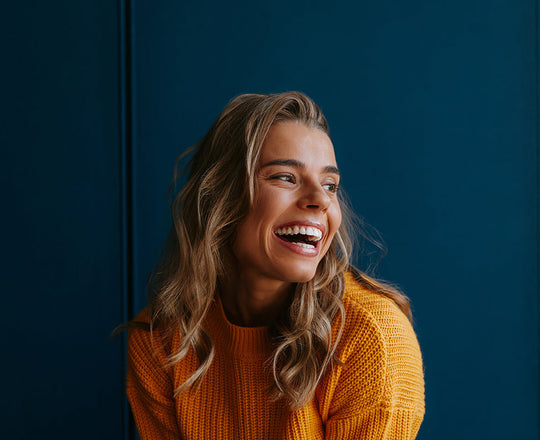 Frau mit gelbem Pullover sitzt vor einer blauen Wand und lächelt