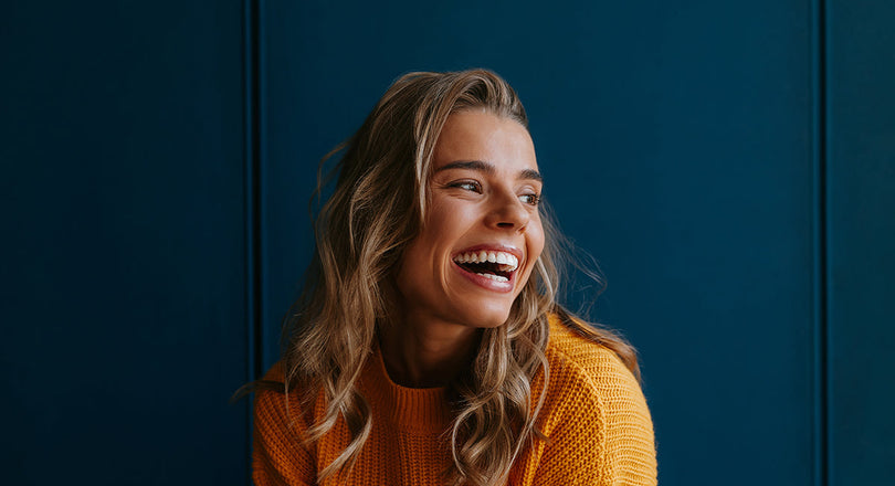 Frau mit gelbem Pullover sitzt vor einer blauen Wand und lächelt