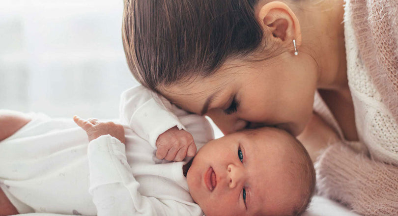 Ein Baby liegt auf einer Decke, während die Mutter ihm einen Kuss auf den Kopf gibt