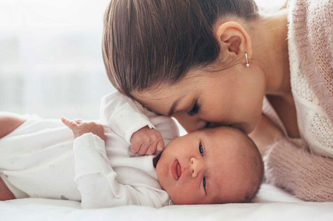 Ein Baby liegt auf einer Decke, während die Mutter ihm einen Kuss auf den Kopf gibt