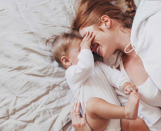 Mama liegt mit ihrem Baby im Bett, das ihr die Augen zuhält