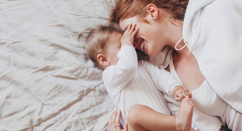 Mama liegt mit ihrem Baby im Bett, das ihr die Augen zuhält