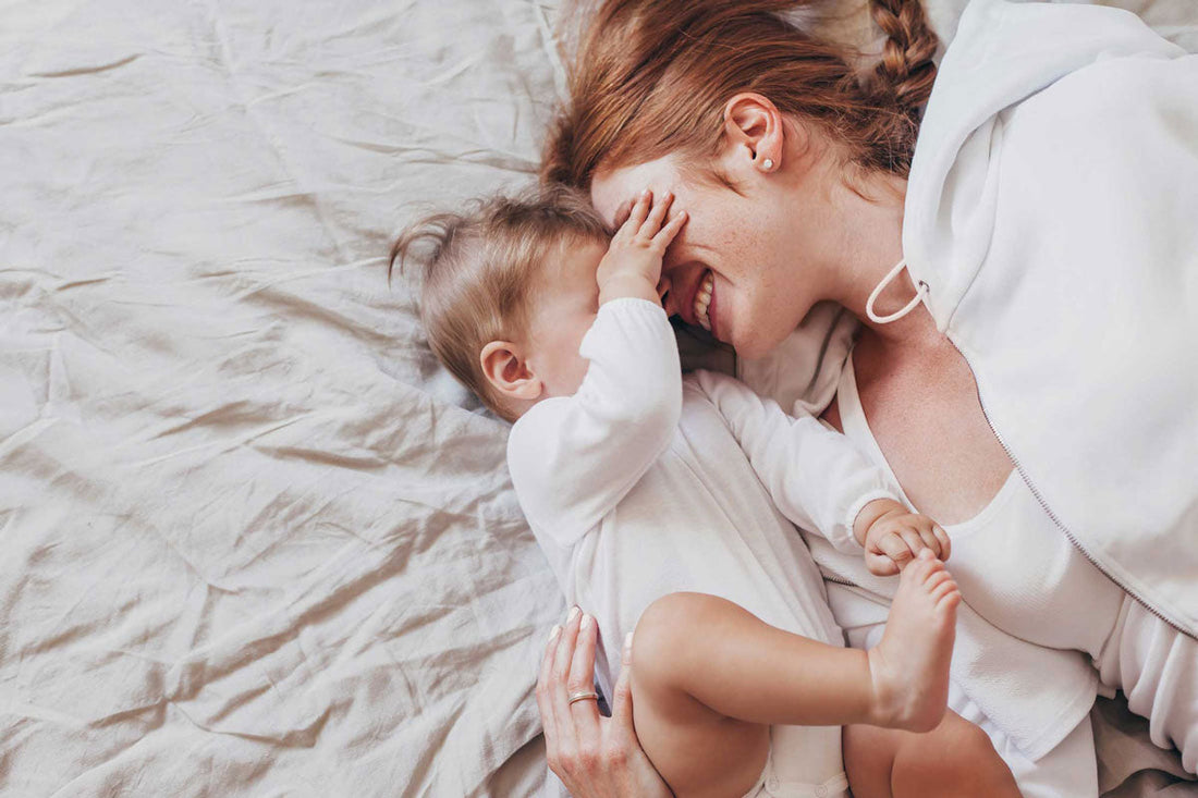 Mama liegt mit ihrem Baby im Bett, das ihr die Augen zuhält