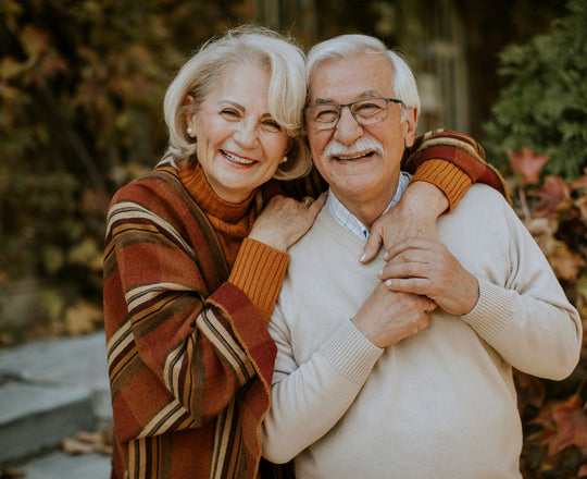 Eine ältere Frau und ein älterer Mann umarmen sich und lächeln vor einem herbstlichen Hintergrund in die Kamera