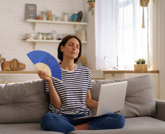 Frau sitzt mit dem Laptop am Bein auf der Couch und hat einen Fächer in der Hand