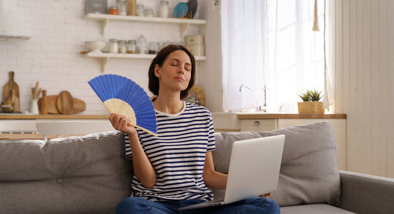 Frau sitzt mit dem Laptop am Bein auf der Couch und hat einen Fächer in der Hand