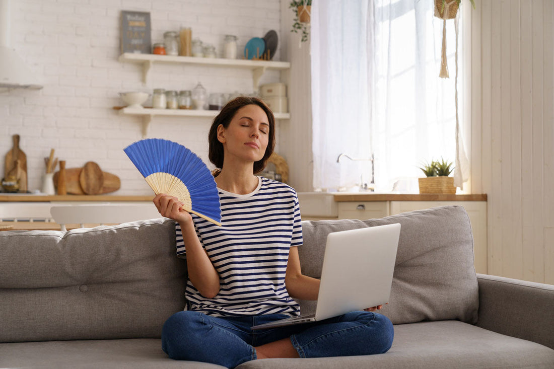 Frau sitzt mit dem Laptop am Bein auf der Couch und hat einen Fächer in der Hand