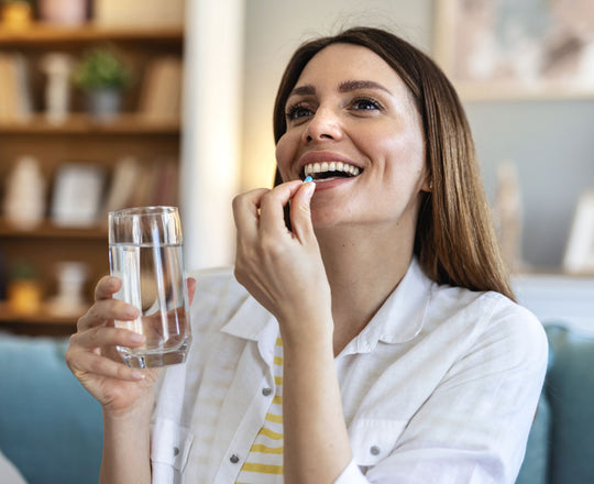 Frau sitzt auf der Couch, hält ein Glas Wasser in der Hand und nimmt eine Kapsel in den Mund