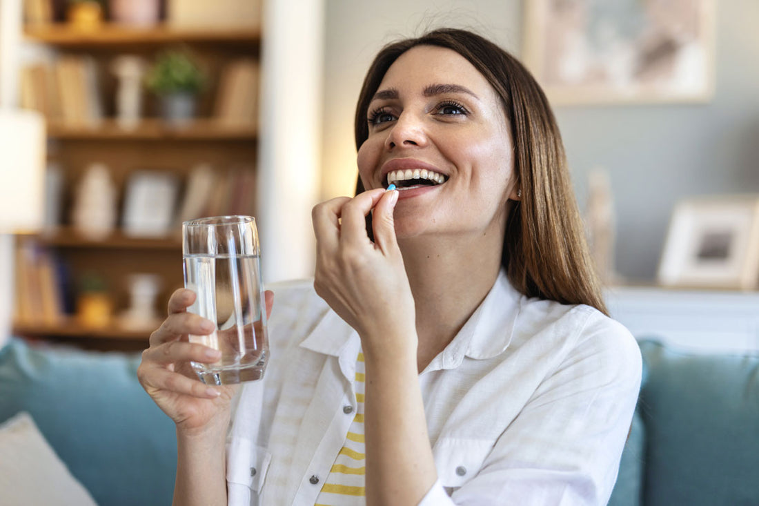 Frau sitzt auf der Couch, hält ein Glas Wasser in der Hand und nimmt eine Kapsel in den Mund