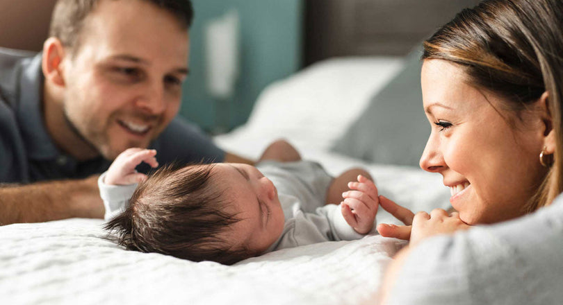 Ein Baby liegt auf einem Bett, während Mama und Papa daneben sitzen und es liebevoll ansehen
