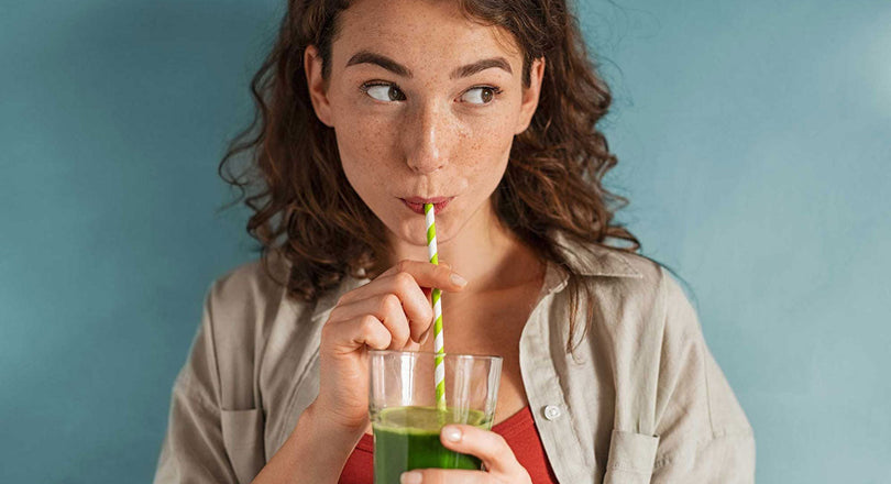 Frau steht vor einer blauen Wand und trinkt einen grünen Smoothie