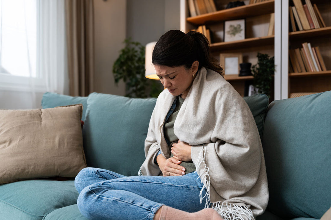 Frau hält sich mit beiden Händen den schmerzenden Bauch, sitzt im Wohnzimmer auf dem Sofa