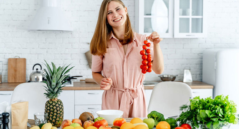Frau steht in der Küche, vor ihr liegt eine Auswahl an buntem Obst und Gemüse