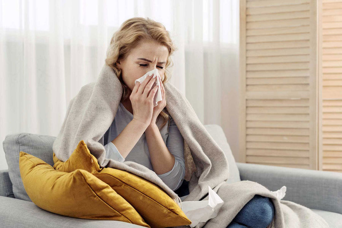 Eine Frau sitzt mit einer Decke auf dem Sofa und schnäuzt sich die Nase