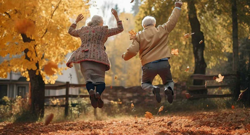Ältere Frau und älterer Mann springen gemeinsam fröhlich in die Luft, umgeben von buntem Herbstlaub