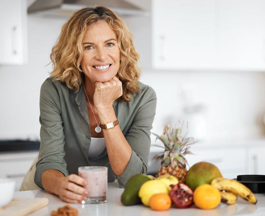Eine Frau lehnt an der Küchentheke, vor ihr stehen ein Glas Smoothie und frisches Obst