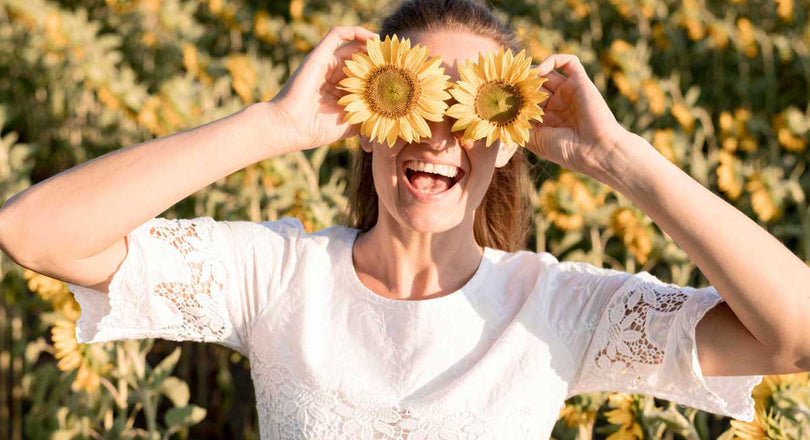 Eine Frau steht vor einem Sonnenblumenfeld und hält zwei Sonnenblumen vor ihre Augen