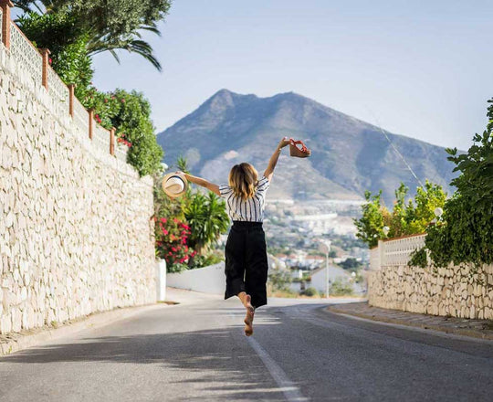 Frau springt in die Luft und hält Sandalen und einen Strohhut in den Händen